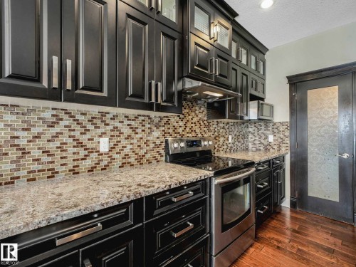 Kitchen featuring dark cabinetry, lighter countertops, a mosaic tile backsplash, stainless steel appliances, and wood-finish flooring - 16251 135 Street Nw, Edmonton, AB - Indoor Photo Showing Kitchen With Upgraded Kitchen