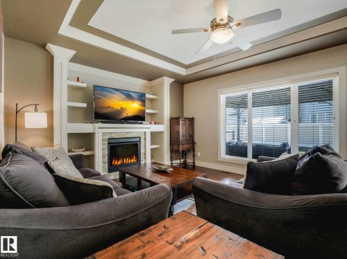 Living space featuring a stone-surround fireplace with built-in shelving, tray ceiling, ceiling fan, and large window with blinds - 16251 135 Street Nw, Edmonton, AB - Indoor Photo Showing Living Room With Fireplace