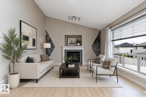 Living area featuring a fireplace with a white mantel and dark tile surround, vaulted ceilings, wood-finish flooring, a large window with blinds, and recessed lighting - 15109 Miller Boulevard, Edmonton, AB - Indoor Photo Showing Living Room With Fireplace