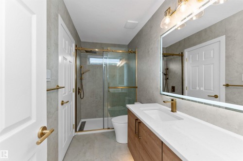 Contemporary bathroom featuring a wood-finish vanity with an integrated white sink and gold-tone faucet, complemented by a backlit mirror and overhead light fixture - 607 Butterworth Wynd, Edmonton, AB - Indoor Photo Showing Bathroom
