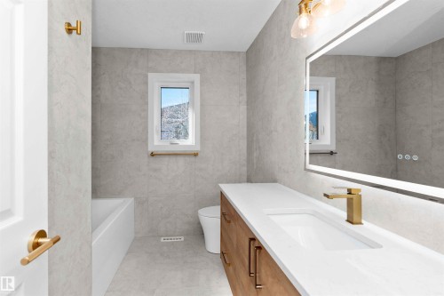 Modern bathroom featuring a single floating vanity with a white countertop and integrated sink, brushed gold hardware, an illuminated mirror, and light gray tiled walls and flooring - 607 Butterworth Wynd, Edmonton, AB - Indoor Photo Showing Bathroom
