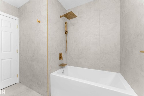 Bathtub and shower combination featuring floor-to-ceiling light-toned wall tiles, matte gold fixtures including a rainfall showerhead, and a white paneled door - 607 Butterworth Wynd, Edmonton, AB - Indoor Photo Showing Bathroom