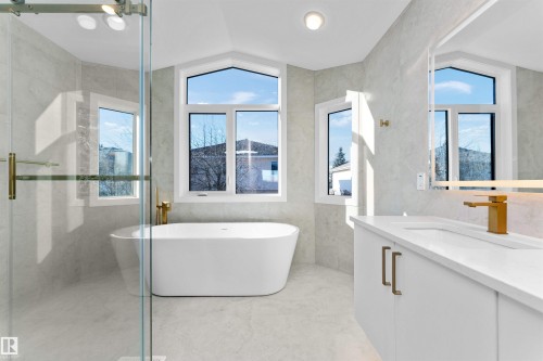 Bathroom featuring a freestanding white bathtub, glass-enclosed shower with gold-tone hardware, white vanity with integrated sink, gold-tone faucet, and light-toned tile flooring and walls - 607 Butterworth Wynd, Edmonton, AB - Indoor Photo Showing Bathroom