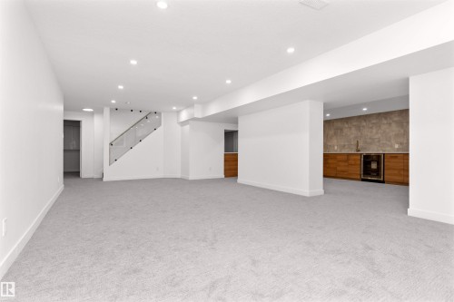 Expansive recreation area featuring light grey carpeting, recessed lighting, a modern glass-paneled staircase, a wet bar with wood-finish cabinetry, and a beverage refrigerator - 607 Butterworth Wynd, Edmonton, AB - Indoor Photo Showing Other Room