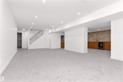 Expansive recreation area featuring light grey carpeting, recessed lighting, a modern glass-paneled staircase, a wet bar with wood-finish cabinetry, and a beverage refrigerator - 