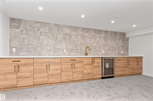 Modern wet bar featuring wood-finish cabinetry, a large basin sink with a gold-tone faucet, a built-in beverage cooler, and a full-height tiled backsplash - 607 Butterworth Wynd, Edmonton, AB - Indoor Photo Showing Other Room