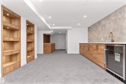 Spacious entertainment area featuring two built-in wood-finish shelving units with integrated lighting, a wet bar with a white countertop and gold-tone faucet, a built-in beverage cooler, and light gray carpeting - 607 Butterworth Wynd, Edmonton, AB - Indoor Photo Showing Other Room