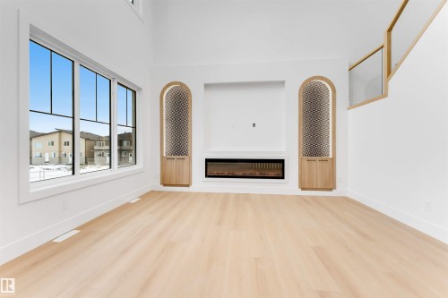This living space features light-toned flooring, a large window with multiple panes, and a modern fireplace integrated into a white wall - 4004 35 Street, Beaumont, AB - Indoor With Fireplace