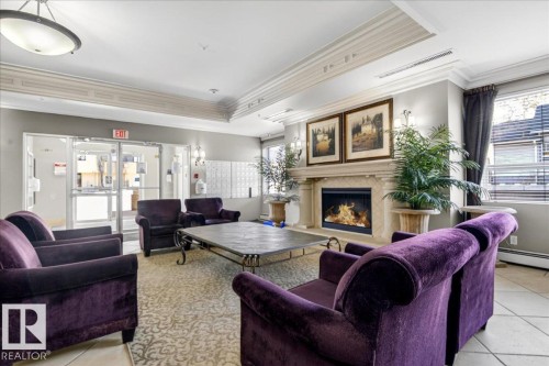 Lobby area featuring a decorative fireplace with a stone mantel, glass entry doors, ceiling-mounted lighting, wall sconces, and crown molding - 701 9707 105 Street, Edmonton, AB - Indoor Photo Showing Living Room With Fireplace