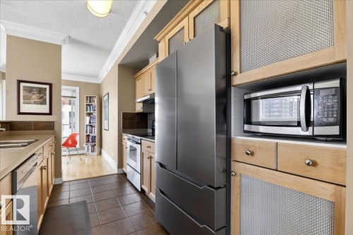 Galley kitchen featuring stainless steel appliances, light wood-finish cabinetry with mesh inserts, and durable tile flooring - 701 9707 105 Street, Edmonton, AB - Indoor Photo Showing Kitchen With Stainless Steel Kitchen With Double Sink