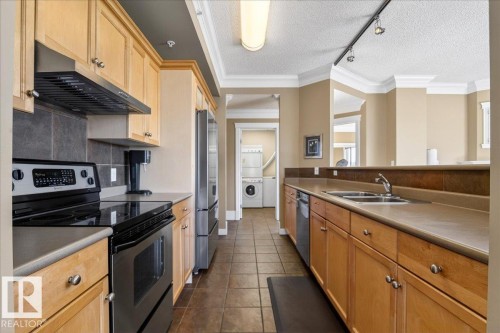 Galley kitchen featuring wood cabinetry, stone-look countertops, stainless steel appliances, and a tile backsplash - 701 9707 105 Street, Edmonton, AB - Indoor Photo Showing Kitchen With Stainless Steel Kitchen With Double Sink