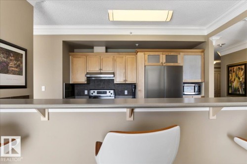 Kitchen featuring light wood cabinetry, stainless steel appliances, dark tile backsplash, a range hood, and a breakfast bar with an overhang - 701 9707 105 Street, Edmonton, AB - Indoor Photo Showing Kitchen