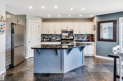 Kitchen featuring a central island with a dark countertop and light blue base, light-colored upper and lower cabinetry, a dark-toned backsplash, stainless steel appliances, and dark-toned slate tile flooring - 96 Bonin Crescent, Beaumont, AB - Indoor Photo Showing Kitchen With Upgraded Kitchen