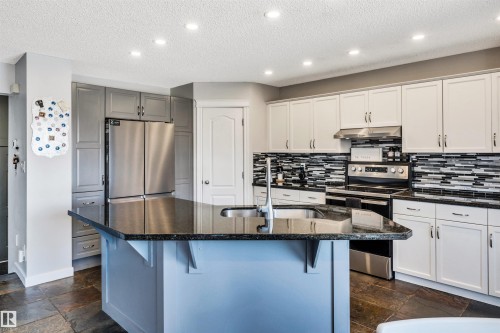 Kitchen featuring a central island with a dark countertop and undermount sink, white cabinetry with silver-tone hardware, a stainless steel electric range with an overhead vent, a mosaic tile backsplash, and slate flooring - 96 Bonin Crescent, Beaumont, AB - Indoor Photo Showing Kitchen With Upgraded Kitchen