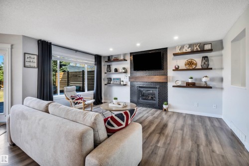 Spacious living area featuring wood-finish flooring, a contemporary fireplace with honeycomb tile surround and wood mantel, large window with natural light, recessed lighting, and wall-mounted shelving - 96 Bonin Crescent, Beaumont, AB - Indoor Photo Showing Living Room With Fireplace