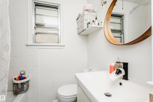 Bathroom featuring a white vanity with an integrated sink, a rectangular window with frosted lower panes, a round mirror with a wood-finish frame, and white subway tile surrounding the tub - 9760 64 Avenue, Edmonton, AB - Indoor Photo Showing Bathroom
