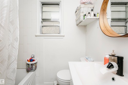 White-tiled bathroom featuring a lavatory with a single-handle faucet, a wall-mounted shelving unit, and a window with privacy glass - 9760 64 Avenue, Edmonton, AB - Indoor Photo Showing Bathroom