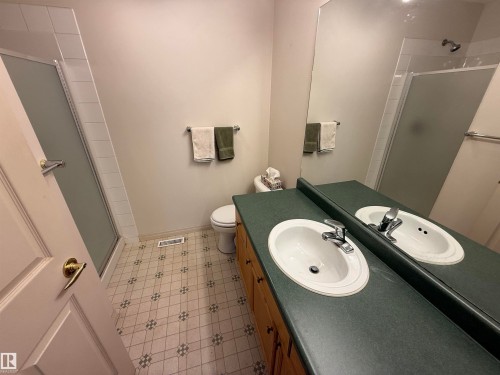 Bathroom featuring a full vanity with dual integrated sinks, an expansive wall mirror, a toilet, and a glass-enclosed shower with tile surround - 14 330 Galbraith Close, Edmonton, AB - Indoor Photo Showing Bathroom