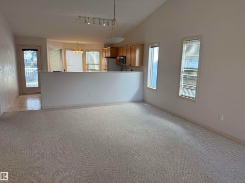 Open concept living area featuring vaulted ceilings, light-toned carpeting, two vertical windows with blinds, a partial wall separating living and kitchen areas, and a door leading to an exterior balcony with white railing - 14 330 Galbraith Close, Edmonton, AB - Indoor