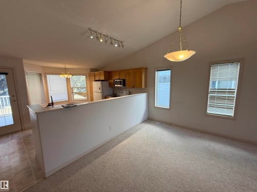 Vaulted ceiling living area featuring neutral carpeting, two windows with blinds, a brass-finish pendant light fixture, and a track lighting fixture - 14 330 Galbraith Close, Edmonton, AB - Indoor