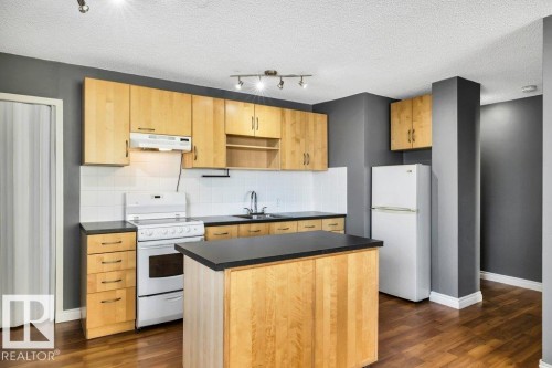 Kitchen featuring wood-finish cabinetry, dark countertops, white subway tile backsplash, a central island, and wood-finish flooring - 403 10021 116 Street, Edmonton, AB - Indoor Photo Showing Kitchen