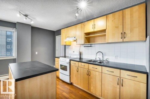 Kitchen featuring light wood cabinetry, dark countertops, a built-in kitchen island, white tile backsplash, and wood-finish flooring - 403 10021 116 Street, Edmonton, AB - Indoor Photo Showing Kitchen With Double Sink