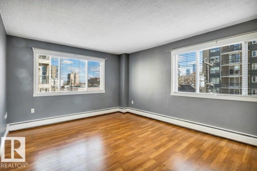 Spacious room with wood-finish flooring, two large windows with blinds, painted walls, white trim, and baseboard heating - 403 10021 116 Street, Edmonton, AB - Indoor Photo Showing Other Room