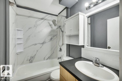 Bathroom featuring a white bathtub with a faux marble surround, a corner shower organizer, a chrome showerhead with an attached handheld sprayer, and a white vanity cabinet with an open shelf - 403 10021 116 Street, Edmonton, AB - Indoor Photo Showing Bathroom