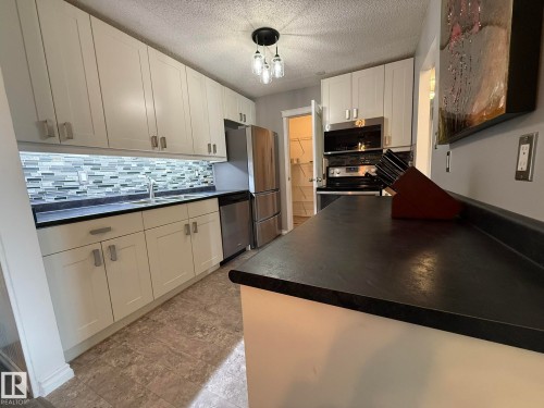 Kitchen featuring white shaker-style cabinetry, dark countertops, a tiled backsplash with varied gray and white tones, stainless steel appliances, and a pantry with wire shelving - 5 2204 118 Street, Edmonton, AB - Indoor Photo Showing Kitchen