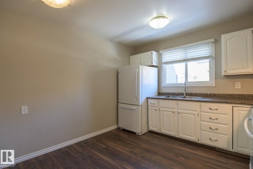 Kitchen featuring wood-finish flooring, white cabinetry with silver hardware, a stainless steel sink with a gooseneck faucet, a white refrigerator, and a window with horizontal blinds - 8039 27 Avenue, Edmonton, AB - Indoor Photo Showing Kitchen With Double Sink