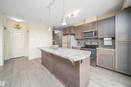 Open-concept kitchen featuring a central island with a granite-style countertop, stainless steel appliances, wood-finish cabinetry, a mosaic tile backsplash, and wood-finish flooring - 421 4008 Savaryn Drive, Edmonton, AB - Indoor Photo Showing Kitchen With Upgraded Kitchen