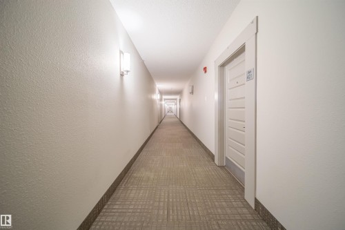 Long hallway featuring recessed ceiling lighting, wall-mounted sconces, textured wall finishes, a pattern-textured carpet, and baseboard trim - 421 4008 Savaryn Drive, Edmonton, AB - Indoor Photo Showing Other Room
