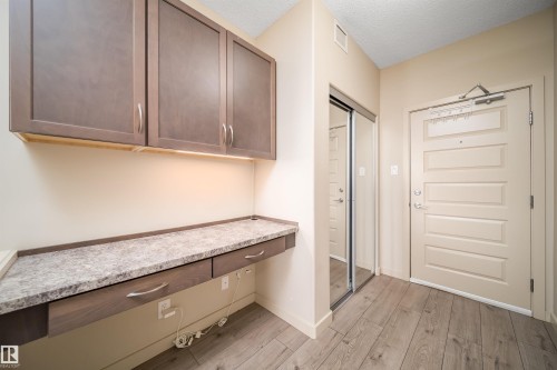 Entryway featuring wood-finish flooring, a built-in desk with two drawers and an integrated countertop, upper cabinetry with under-cabinet lighting, a mirrored sliding closet door, and a paneled entry door - 421 4008 Savaryn Drive, Edmonton, AB - Indoor Photo Showing Other Room