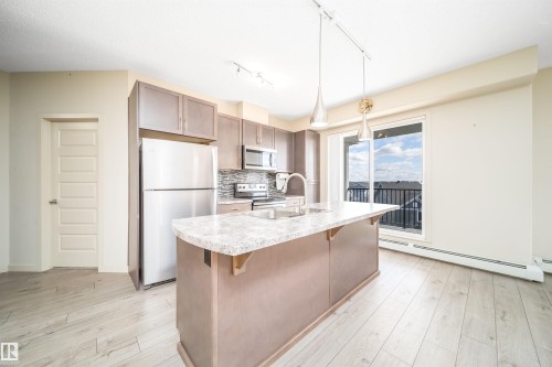 Modern kitchen featuring an island with composite countertops, stainless steel appliances, wood-finish flooring, and a sliding glass door to an outdoor space - 421 4008 Savaryn Drive, Edmonton, AB - Indoor Photo Showing Kitchen