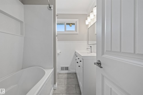 Contemporary bathroom featuring a white vanity with a single sink, an integrated bathtub with a shower niche, a window offering natural light, modern lighting fixtures, and tile flooring - 7927 71 Avenue, Edmonton, AB - Indoor Photo Showing Bathroom