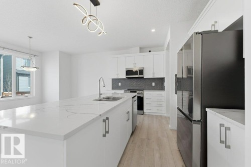 Spacious kitchen featuring an oversized island with a built-in sink, white cabinetry with contemporary hardware, stainless steel appliances, a dark tile backsplash, and light wood-finish flooring - 2011 207 Street, Edmonton, AB - Indoor Photo Showing Kitchen With Double Sink With Upgraded Kitchen