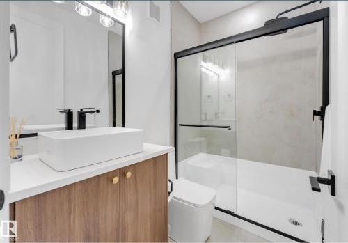 Contemporary bathroom featuring a wood-finish vanity with a vessel sink, a walk-in shower with clear glass enclosure and black trim, a square toilet, and modern wall-mounted lighting - 4727 17 Avenue Sw, Edmonton, AB - Indoor Photo Showing Bathroom