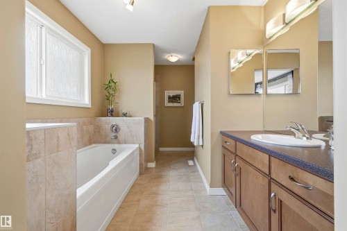 Bathroom featuring a built-in tub with tile surround, a window with obscure glass, and a vanity with wood-finish cabinetry, a white basin, and a wall-mounted mirror - 2340 Hagen Link, Edmonton, AB - Indoor Photo Showing Bathroom