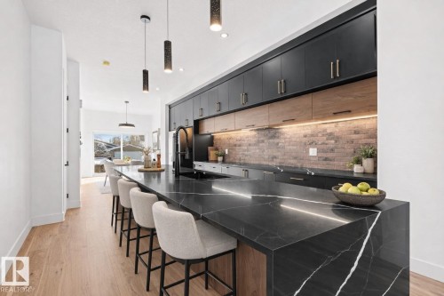 Contemporary kitchen featuring a large black stone island with white veining, undercabinet lighting, dark cabinetry with brass hardware, a dark brick-patterned backsplash, and light wood-finish flooring - 11021 132 Street, Edmonton, AB - Indoor Photo Showing Kitchen With Upgraded Kitchen