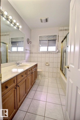 Bathroom featuring a dual-sink vanity with wood cabinetry, a large mirror with strip lighting, tiled flooring, and a shower-tub combination with a glass enclosure - 9512 134 Avenue, Edmonton, AB - Indoor Photo Showing Bathroom