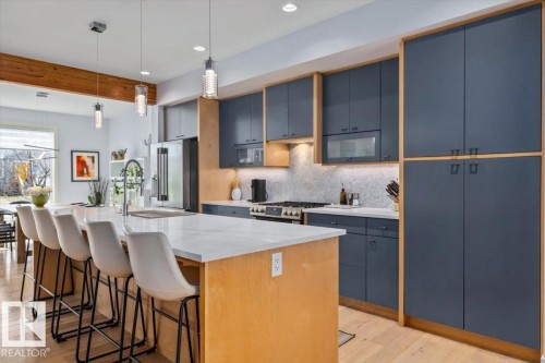 Modern kitchen featuring a large central island with a light-toned countertop, sleek dark cabinetry with wood-finish framing, under-cabinet lighting, a stainless steel range, and wood-finish flooring - 11245 64 Street, Edmonton, AB - Indoor Photo Showing Kitchen With Upgraded Kitchen