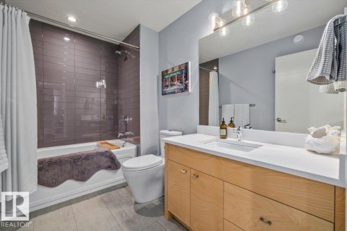 Bathroom featuring a light wood-finish vanity with a white countertop and integrated sink, a rectangular mirror with an overhead multi-bulb light fixture, a white toilet, and a combined tub and shower with gray subway tile surround - 11245 64 Street, Edmonton, AB - Indoor Photo Showing Bathroom