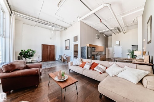 Open-concept living area featuring high ceilings with exposed ductwork, dark wood-finish flooring, and large industrial-style windows - 1003 10179 105 Street Nw, Edmonton, AB - Indoor Photo Showing Living Room