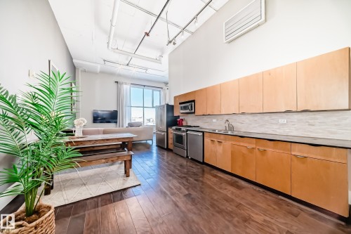 Open-concept living space featuring high ceilings, wood-finish flooring, a full kitchen with light wood cabinetry and stainless steel appliances, and large windows providing ample natural light - 1003 10179 105 Street Nw, Edmonton, AB - Indoor