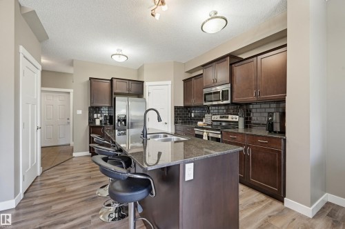 Kitchen featuring a large central island with seating, dark wood-finish cabinetry, stainless steel appliances, dark subway tile backsplash, and wood-finish flooring - 11 94 Longview Drive, Spruce Grove, AB - Indoor Photo Showing Kitchen With Double Sink