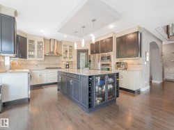 Spacious kitchen featuring a large central island with integrated storage, stainless steel appliances, a range hood, wood-finish flooring, and a mix of dark and light cabinetry with glass-front accents - 