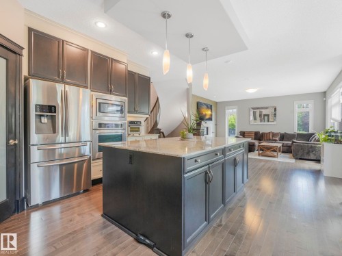 Gourmet kitchen featuring a large island with granite-style countertop, stainless steel appliances, dark wood-finish cabinetry, and recessed lighting, opening to a spacious living area with wood-finish flooring - 932 Summerside Link, Edmonton, AB - Indoor Photo Showing Kitchen With Upgraded Kitchen