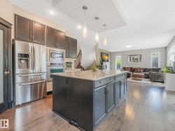 Gourmet kitchen featuring a large island with granite-style countertop, stainless steel appliances, dark wood-finish cabinetry, and recessed lighting, opening to a spacious living area with wood-finish flooring - 