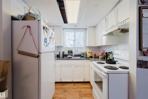Compact kitchen featuring white cabinetry, a white electric range, a white refrigerator, wood-finish flooring, and a window with blinds - 10831 80 Avenue, Edmonton, AB - Indoor Photo Showing Kitchen With Double Sink
