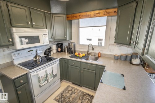 Kitchen featuring painted shaker-style cabinetry, a stainless steel sink with a gooseneck faucet, a white electric range with an overhead microwave, and light-toned countertops - 4417 46A St, St. Paul Town, AB - Indoor Photo Showing Kitchen